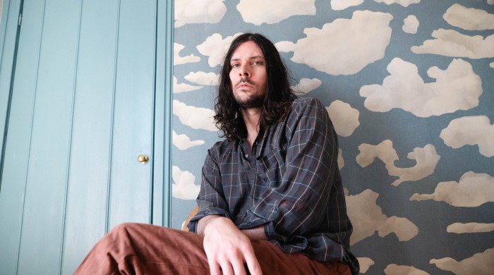 Man sitting against wall with long dark hair, wearing blue check shirt and brown trousers
